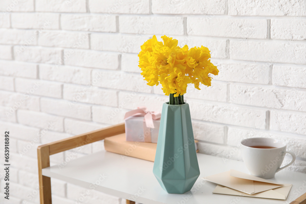 Vase with narcissus flowers, gift, cup of tea and envelopes on table near white brick wall