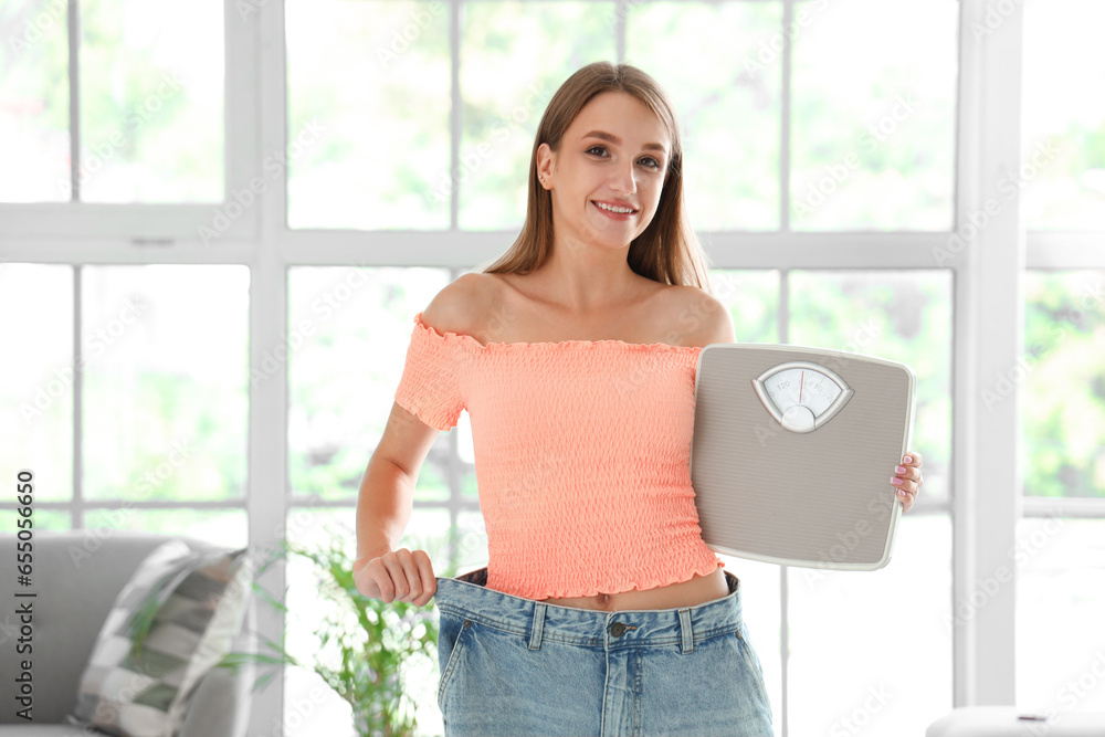 Beautiful young woman in loose jeans with scales at living room. Weight loss concept