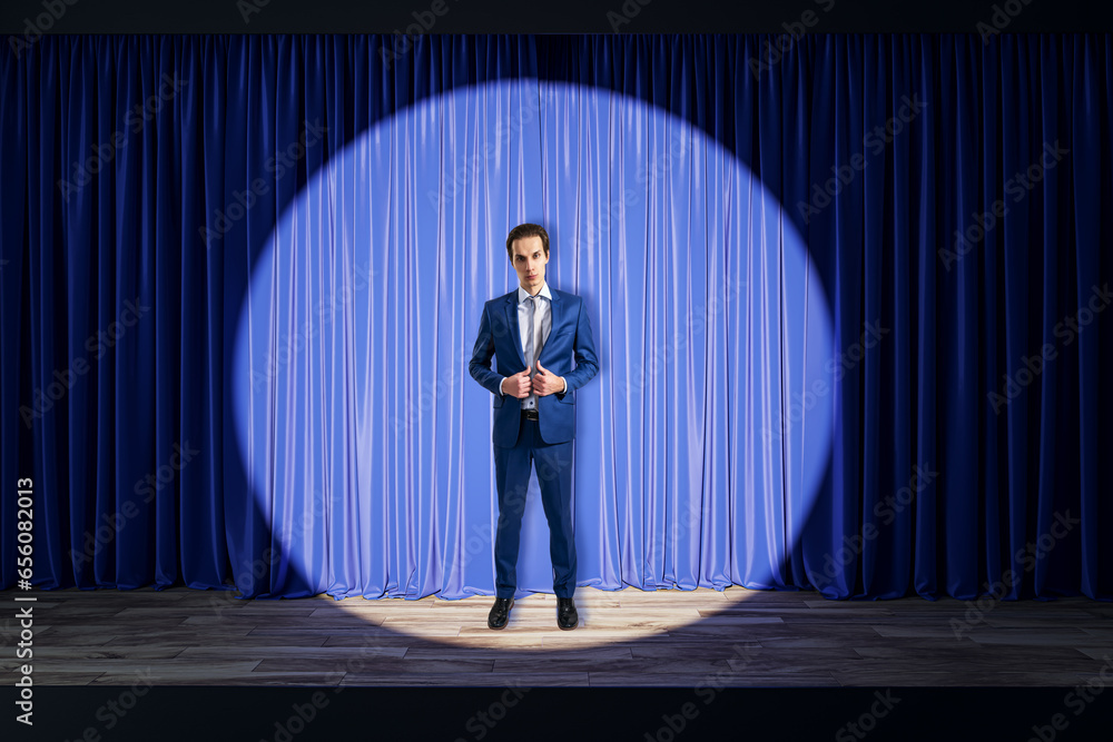 Attractive young businessman standing in front of blue curtains in spotlight in theater. Presentation and speech concept.