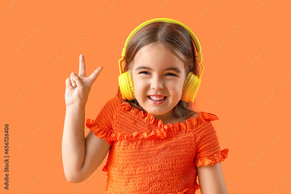 Cute little girl in headphones showing victory gesture on orange background