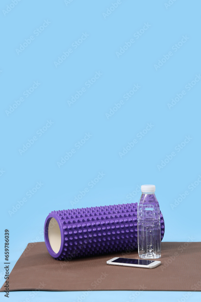 Fitness mat with foam roller, mobile phone and bottle of water on blue background