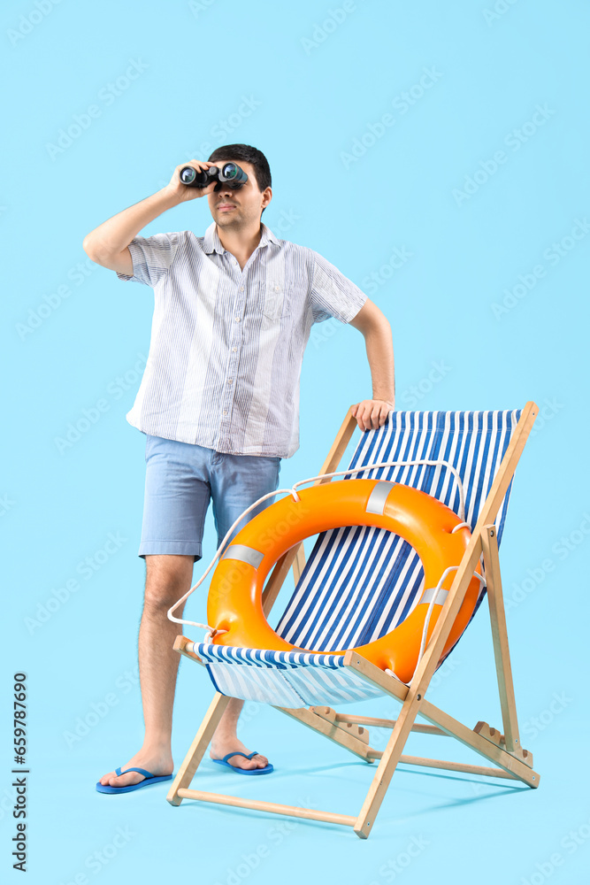 Young man with binoculars, ring buoy and deck chair on blue background