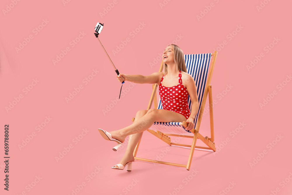 Beautiful young woman taking selfie in deck chair on pink background