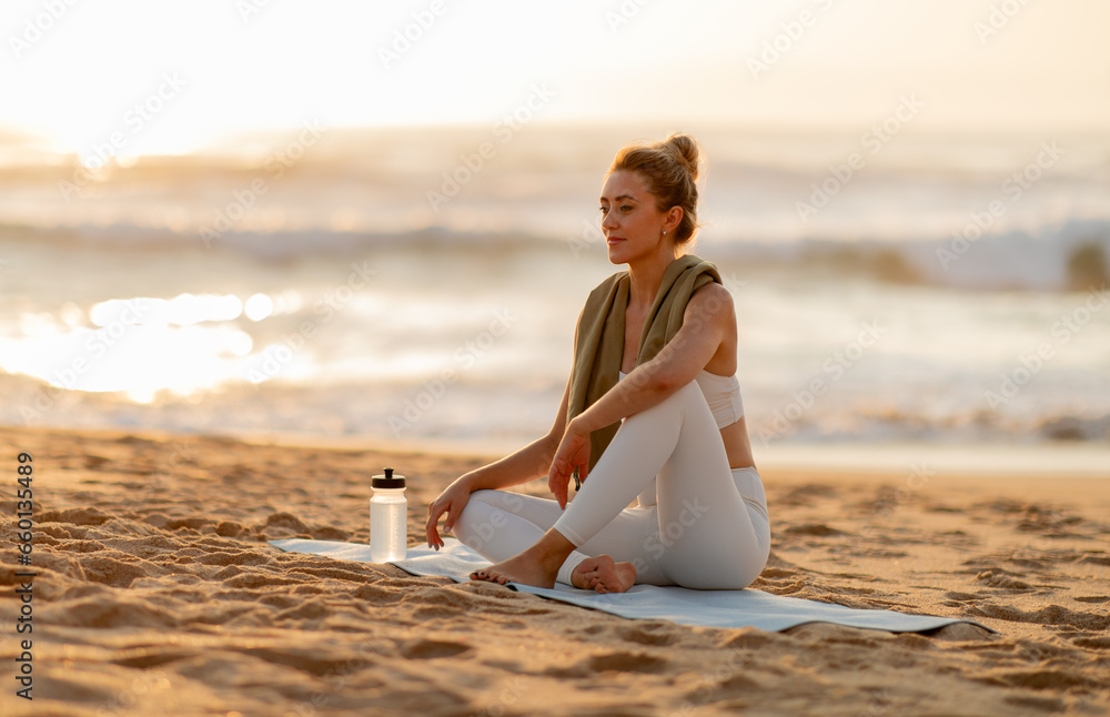 Young woman relaxing after outdoor training, sitting on fitness mat on sea beach and looking away, resting after workout