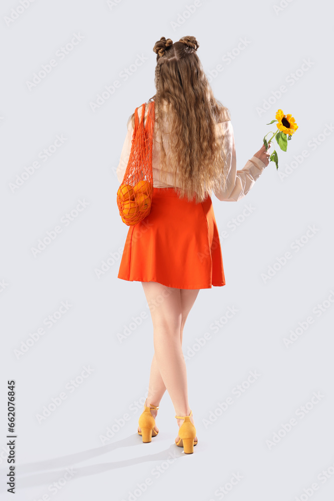 Young woman with beautiful sunflower and string bag on light background, back view