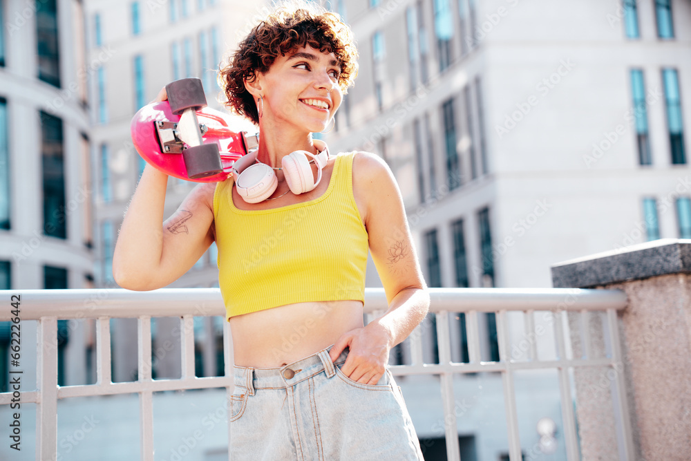 Young beautiful smiling hipster woman in trendy summer clothes. Carefree woman with curls hairstyle, posing in the street. Positive model outdoors. Cheerful and happy. Holds penny skateboard