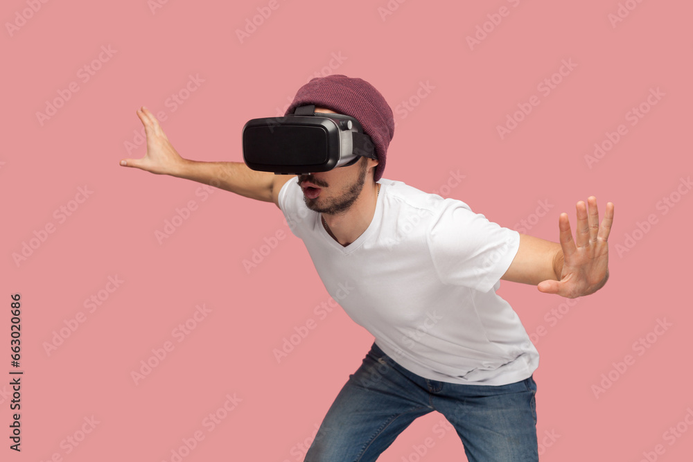Portrait of amazed wary bearded man in white T-shirt and beany hat, wearing vr, playing in headset, trying to avoid monster in video game. Indoor studio shot isolated on pink background.