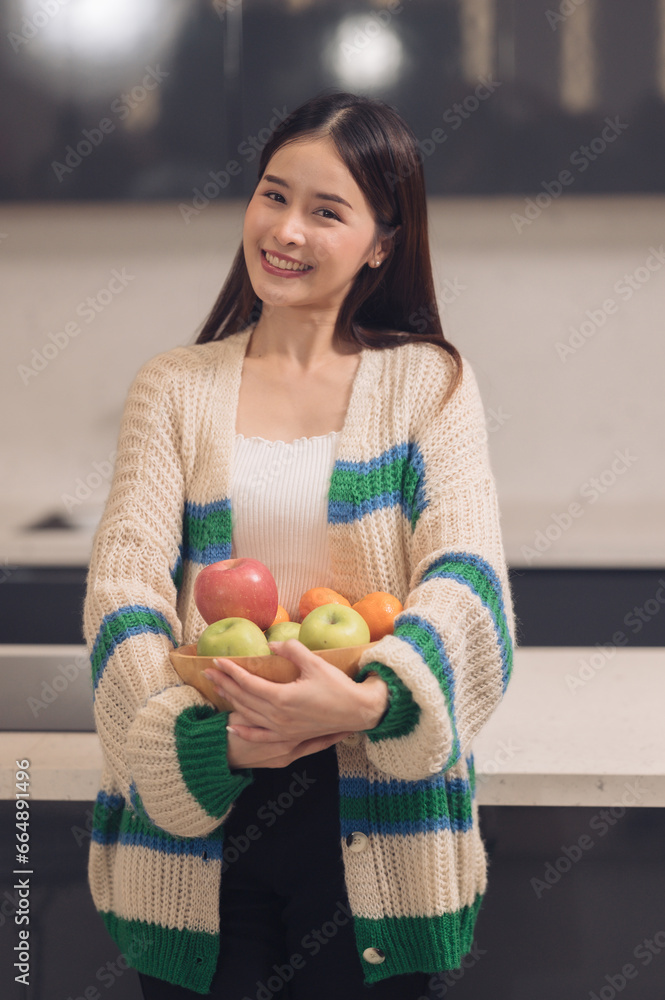 Asian woman wearing a sweater holding fruit with vitamins for good health Standing in the kitchen making breakfast in the morning