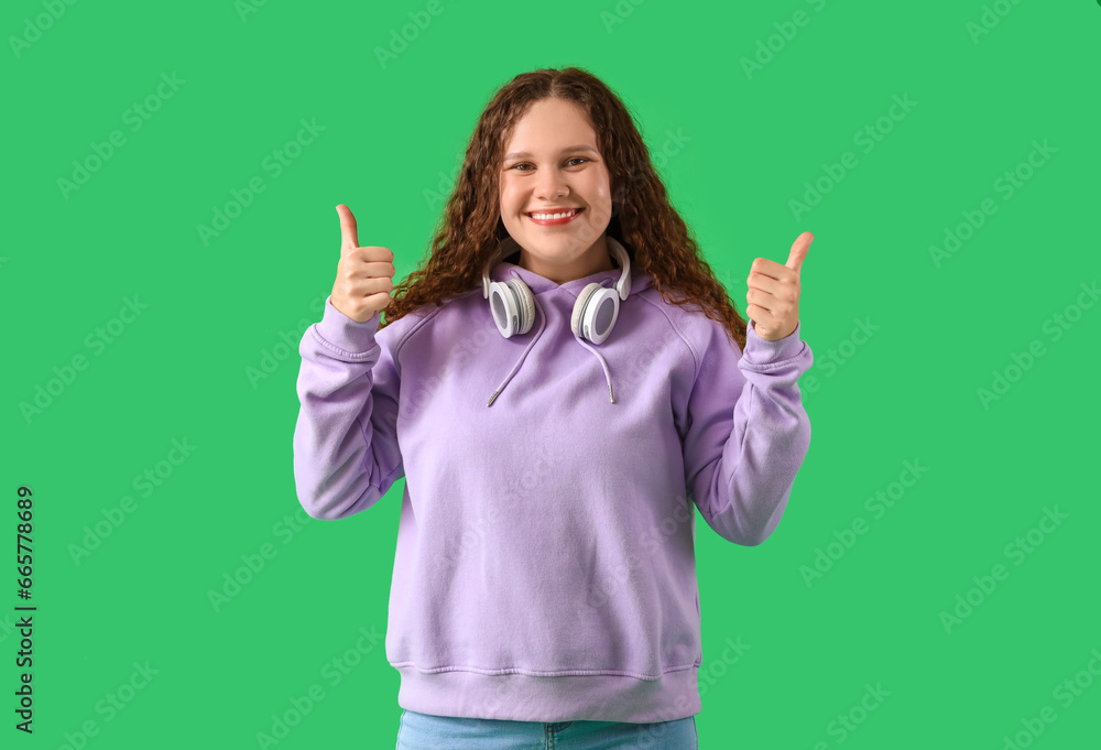 Beautiful young woman in stylish hoodie with headphones showing thumbs-up gesture on green background