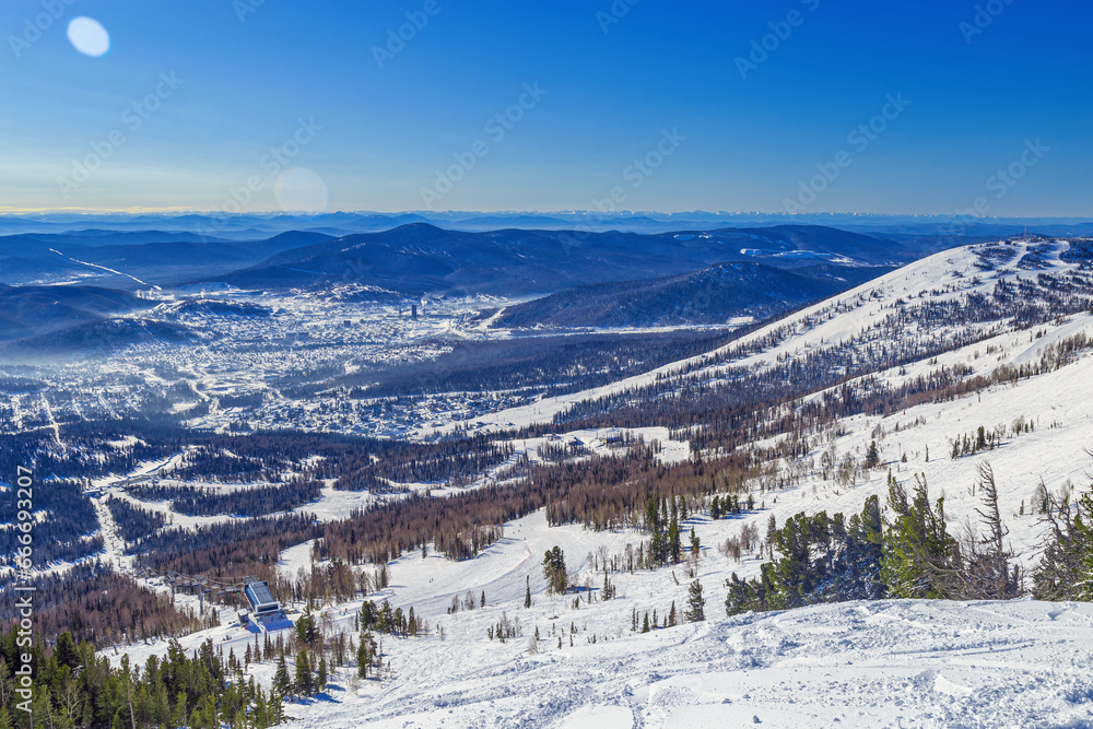 Sheregesh ski resort in Russia, picturesque nature landscape, sun glare on blue sky, white snow slopes and ski trails, view with range mountains and hills, forest, sun and snow, winter weather