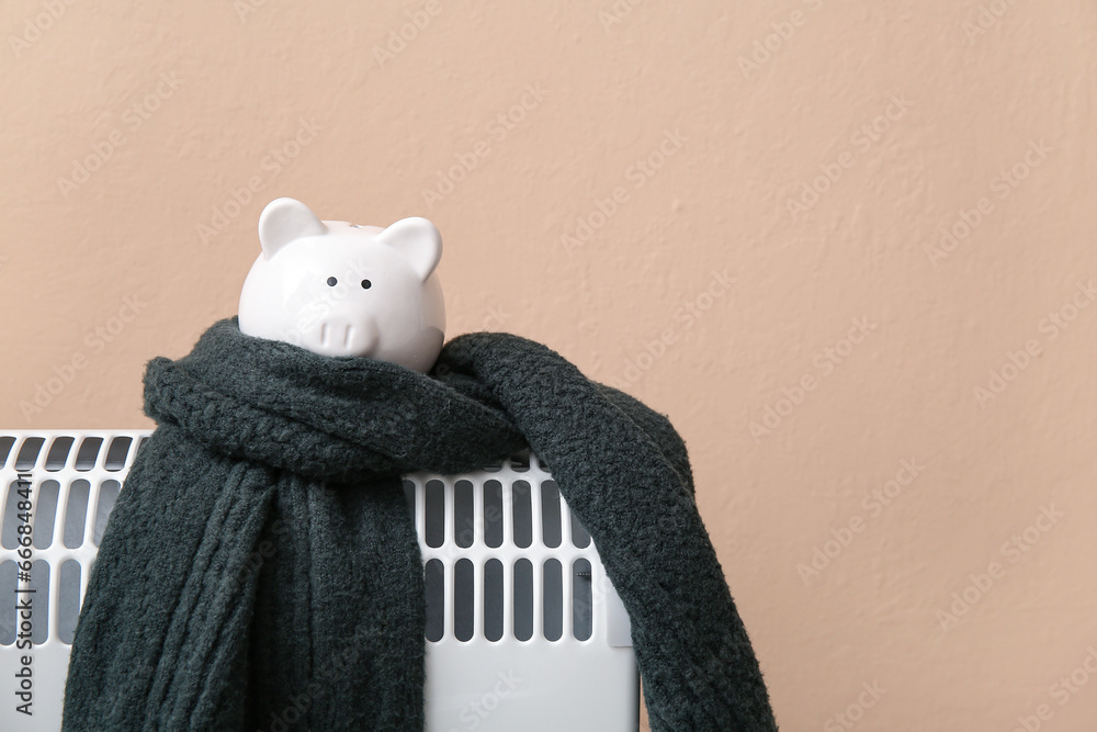 Piggy bank with warm scarf on electric convector heater near beige wall at home. Heating saving concept