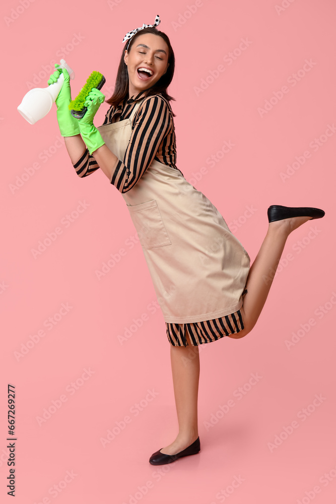 Portrait of happy young housewife in apron with spray bottle and brush on pink background