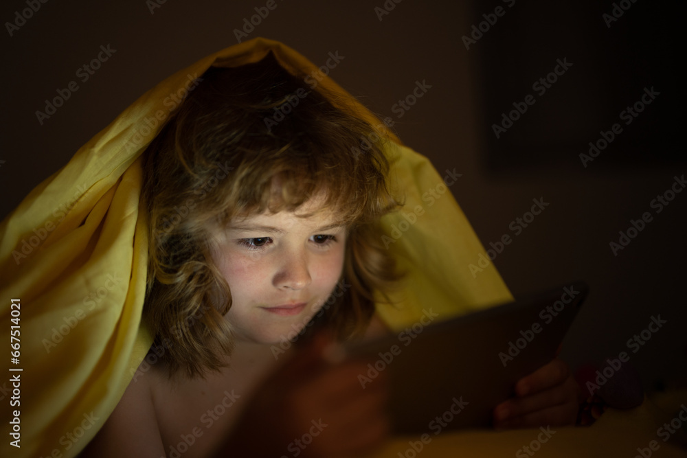 Kid lying in bed at night and playing digital tablet. Child watching tablet. Child in bed at night watching tablet, face illuminated by light of screen. Bed time story.
