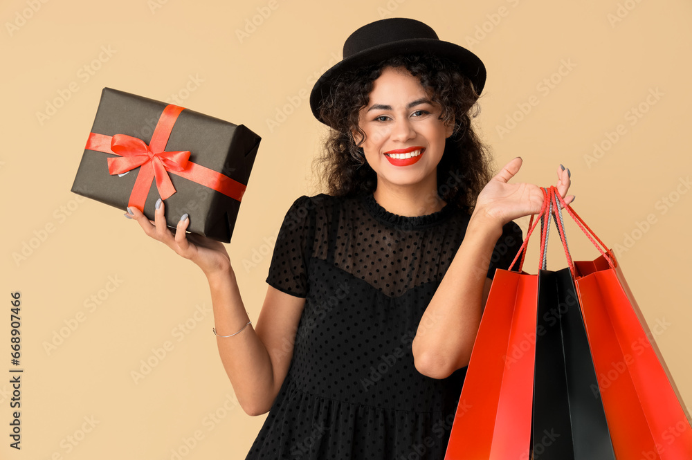 Beautiful African-American woman with gift box and shopping bags on beige background. Black Friday sale