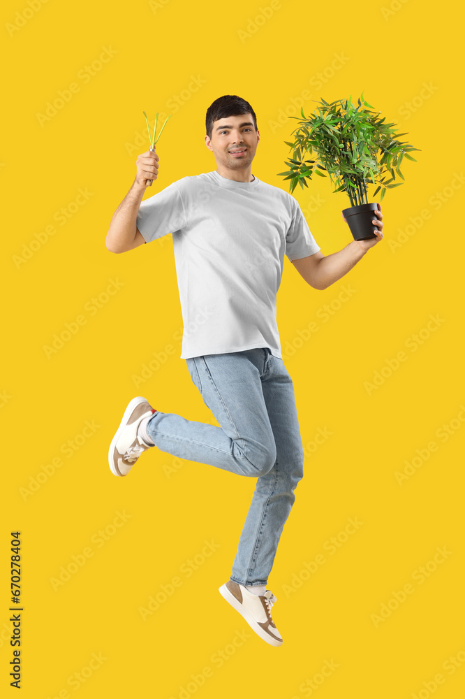 Young man with plant and gardening tool jumping on yellow background