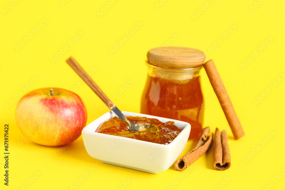 Jar and bowl of sweet apple jam with cinnamon on yellow background