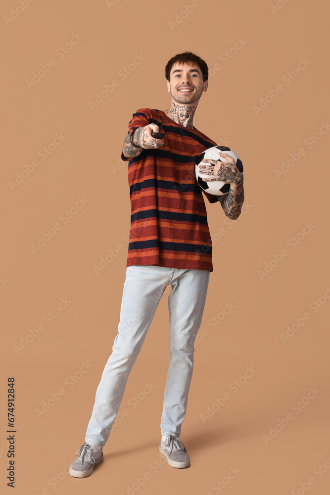 Happy young man with soccer ball and TV remote on brown background