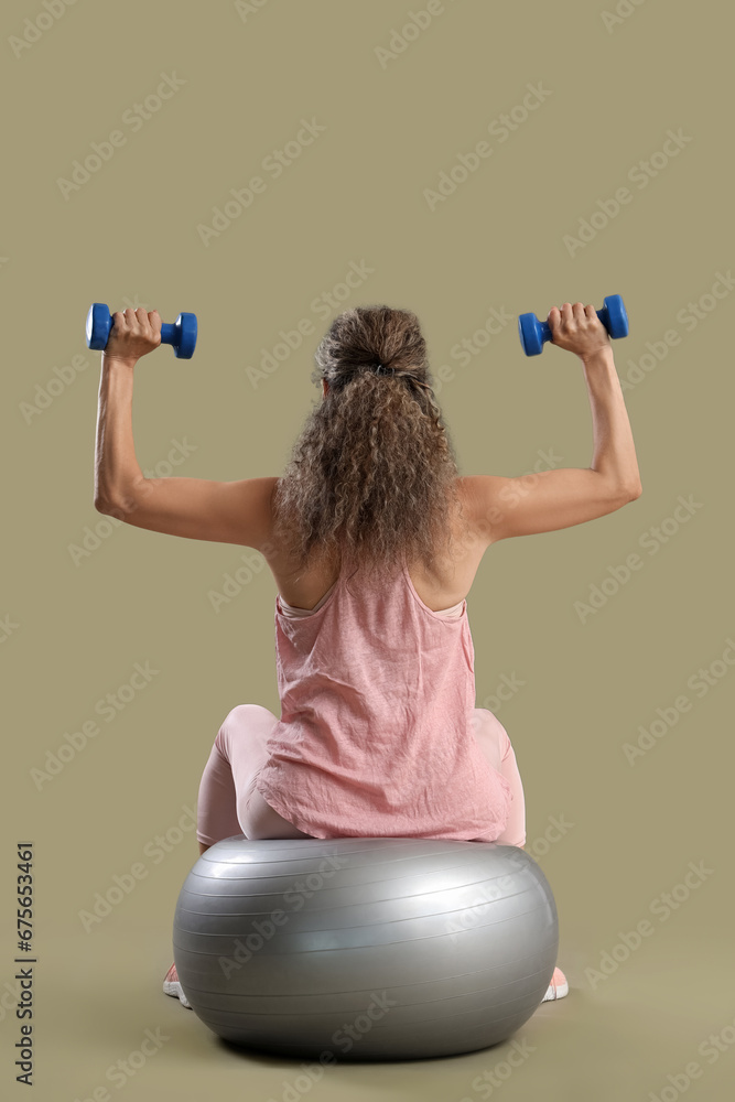 Adult woman exercising with dumbbells on fitness ball against green background, back view