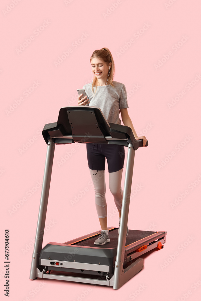 Sporty young woman listening to music on treadmill against pink background