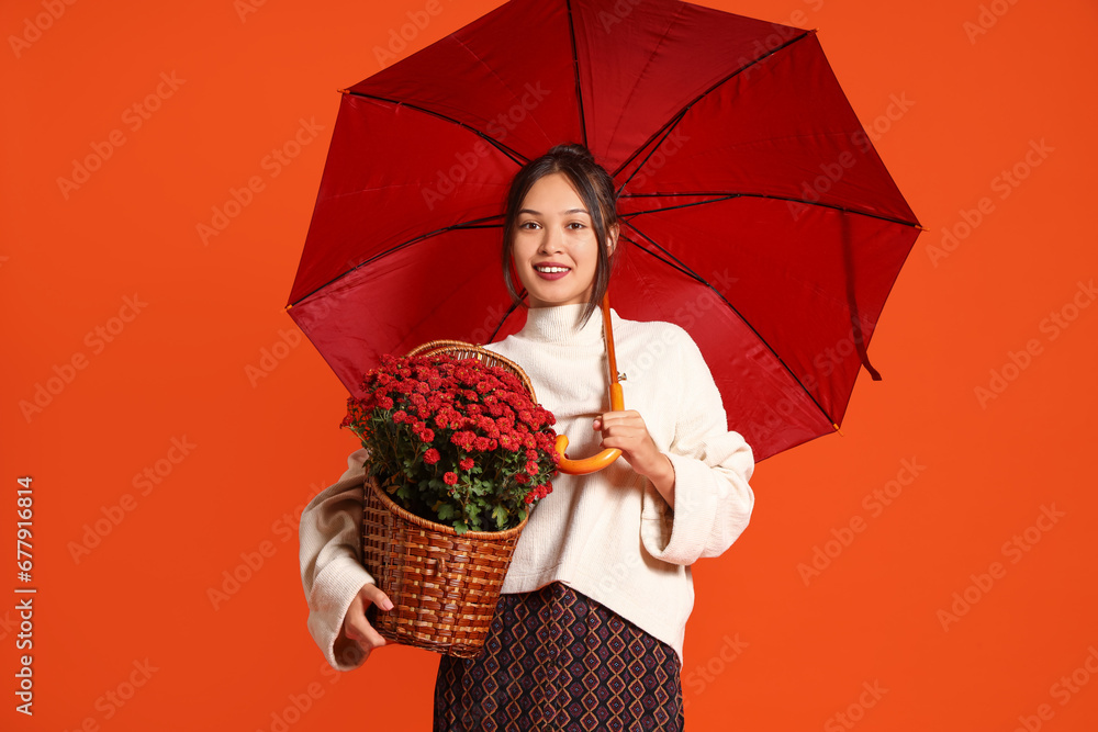 Young Asian woman with umbrella and chrysanthemum flowers on orange background