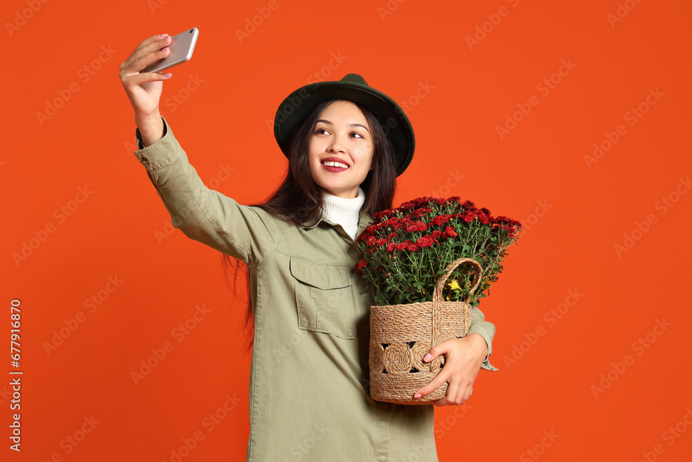 Young Asian woman with chrysanthemum flowers taking selfie on orange background