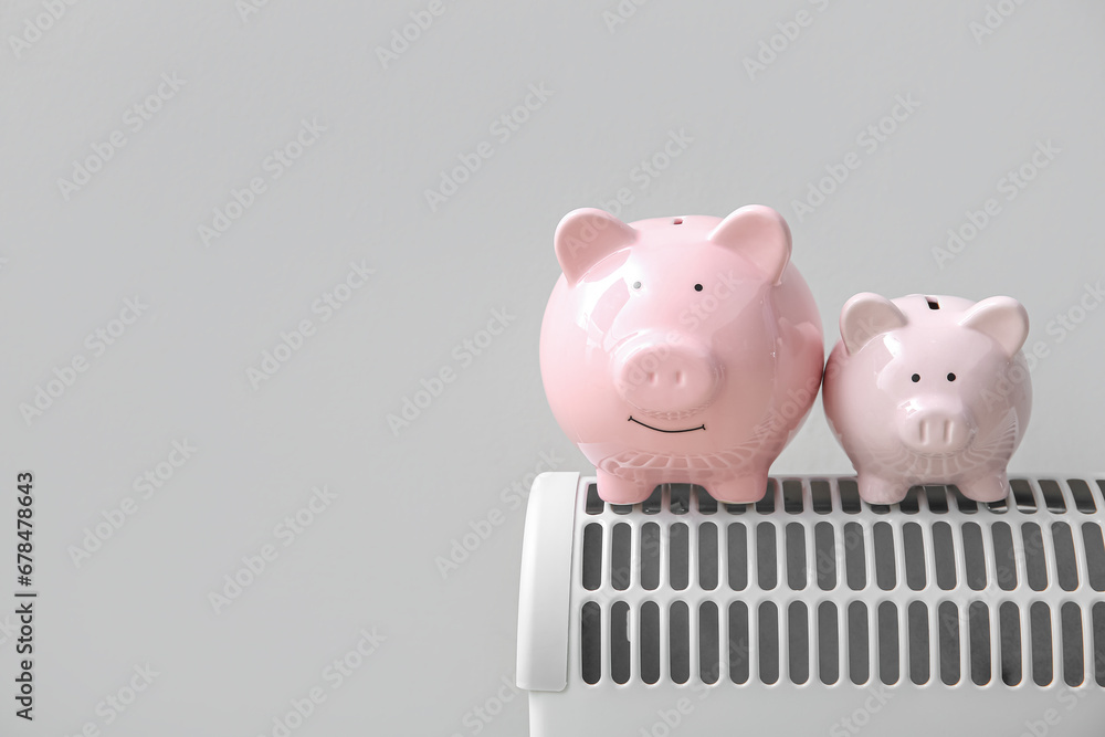 Piggy banks on electric convector heater near white wall at home. Heating saving concept